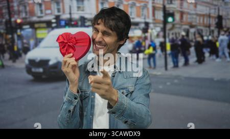 Man holding red heart shaped gift box near smiling face in bustling crowded urban street; love romance. Stock Photo