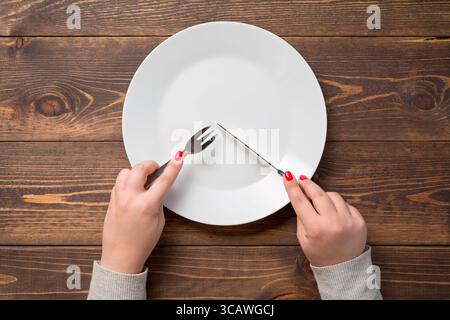 Female hands with knife and fork over white empty plate. Wooden table. Stock Photo