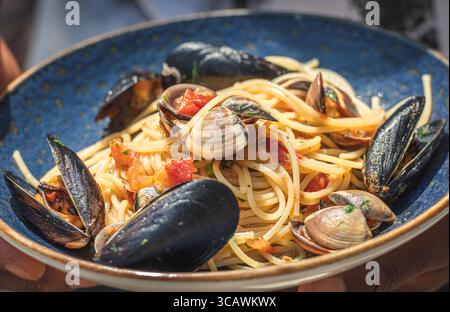 Spaghetti with clams and black mussels, fresh cherry tomatoes, parsley leaves and ground black pepper served in a blue ceramic plate Stock Photo