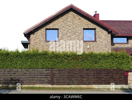 Behind a wooden fence and green coniferous hedge is the side wall of a standard no name yellow rural brick house. Blue sky reflections in windows. Iso Stock Photo