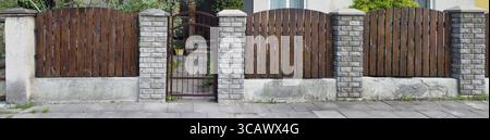 The simple fence of the old no name European house is made of vertical brown thin pine boards. Columns from a decorative white brick. Sunny spring day Stock Photo