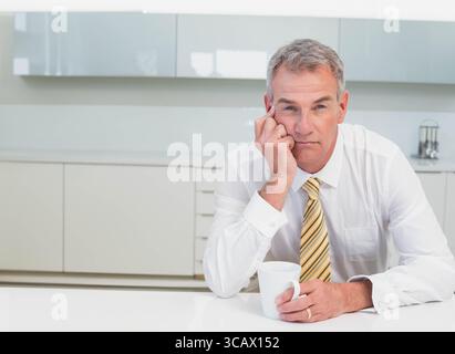 Middle-aged male wearing striped tie leaning on home kitchen counter holding white mug, copy space Stock Photo