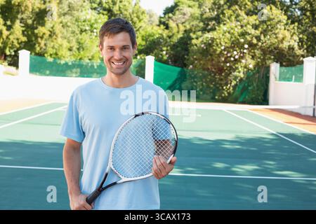 Man smiling and holding tennis racket at chest on sunlit green tennis court by tennis net Stock Photo