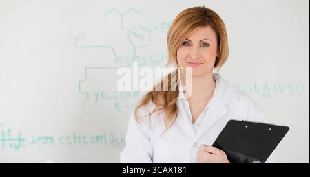 Female scientist standing and holding clipboard by whiteboard with formulas at lab, copy space Stock Photo