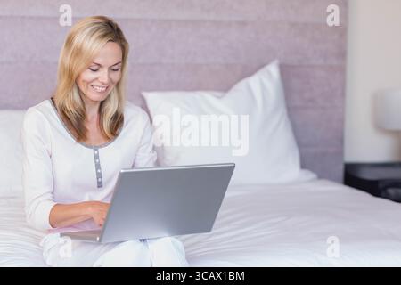 Middle-aged woman sitting cross-legged on bed typing on laptop in bedroom with pillow and lamp Stock Photo