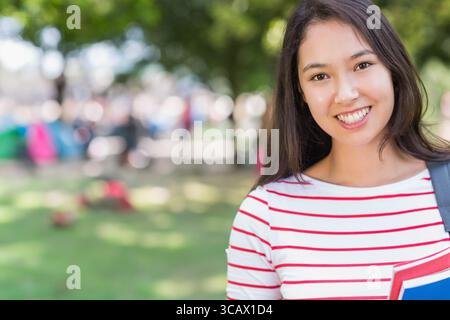 Asian woman student standing on quad carrying red notebook, blue binder and backpack, copy space Stock Photo