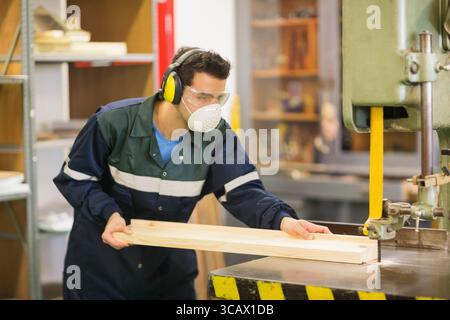 Woodworker operating vertical bandsaw cutting long wooden plank in workshop near metal shelving Stock Photo