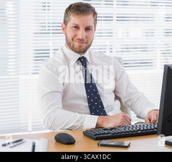 Man wearing white shirt and blue tie typing at office desk with keyboard, monitor and smartphone Stock Photo