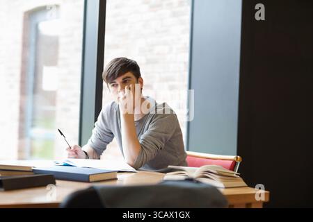 Male student sitting at wooden table by window holding pen over open notebook beside books Stock Photo