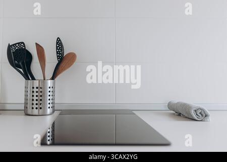 Minimalist kitchen scene with utensil set in metallic holder and black cooktop on clean white surface. Stock Photo