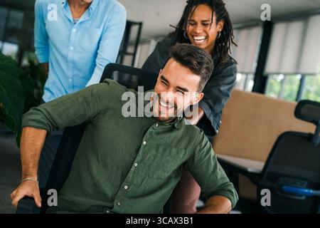 Young colleagues group having fun together riding on chairs in office diverse excited office workers Stock Photo