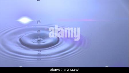 Hovering water droplet creating concentric ripples on calm water surface in studio, with lens flare Stock Photo