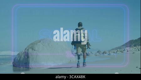 Walking man with prosthetic leg along sandy shoreline, with backpack, board and camera tripod Stock Photo