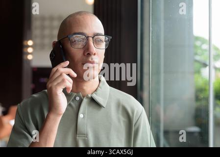Latin adult man in eyeglasses using phone while seated in hotel lobby or bar lounge. Modern digital lifestyle, casual business setting, communication Stock Photo