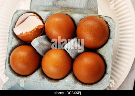 six golden brown chicken eggs in a pale blue egg box with one broken egg Stock Photo