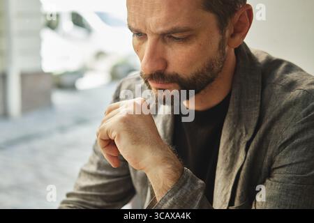 Pensive man in a blazer sitting at a table outdoors, resting his chin on his hand and looking down. Thoughtful, reflective expression Stock Photo