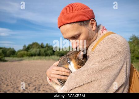 Close up tender man kisses cuddles frightened cat burying face in owner hands during beach walk Stock Photo