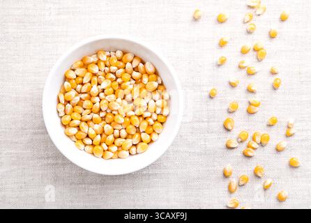 Popcorn kernels, in a white bowl on linen. Seeds of Zea mays variety everta, rounded at the top, which expand and puffs up, when heated. Stock Photo