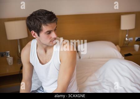 Man sitting on edge of unmade bed in hotel-style bedroom wearing pajamas checking smartphone Stock Photo