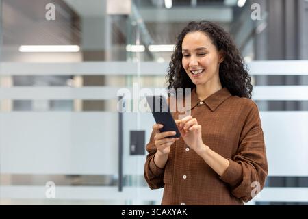 A cheerful woman in a casual brown outfit interacts with her phone in a contemporary office space, evoking connectivity and professionalism. Stock Photo