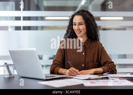 A professional woman sits at a desk, smiling while working on papers and using her laptop. Stock Photo