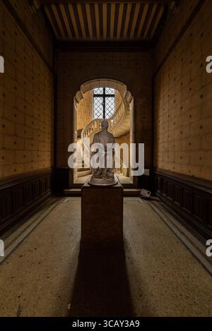 Historic statue illuminated by natural light in an abandoned French chateau with a spiral staircase in the background Stock Photo