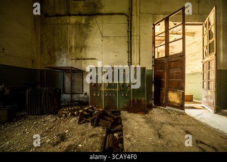 Rusty electrical control panel and broken machinery in an abandoned industrial building with open wooden doors and natural light Stock Photo