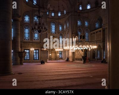 Interior of a historic mosque in Istanbul with worshippers praying on carpeted floor under ornate chandeliers and arched windows Stock Photo
