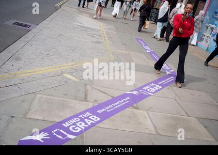 A phone user and other pedestrians walk past pavement messages on Oxford Street that warn them of the increasing trend of phone snatch crime, the 'Mind the Grab' initiative by Westminster, the Met Police and the Currys retailer, on 7th August, 2025, in London, England. Statistics reveal that in 2024, a phone was stolen every 15 minutes in Westminster - 94 a day and over 34,000 in total. Typically, these thefts involve criminals on mopeds or e-bikes snatching phones straight from people’s hands on busy pavements. Stock Photo