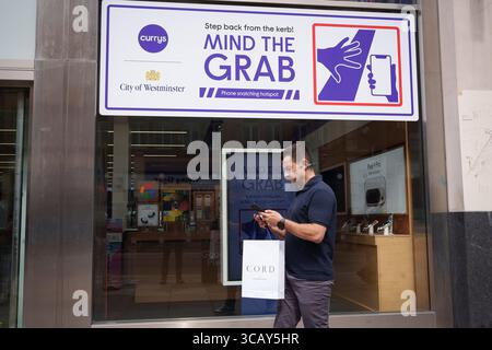 A phone user and other pedestrians walk past messages on Oxford Street that warn them of the increasing trend of phone snatch crime, the 'Mind the Grab' initiative by Westminster, the Met Police and the Currys retailer, on 7th August, 2025, in London, England. Statistics reveal that in 2024, a phone was stolen every 15 minutes in Westminster - 94 a day and over 34,000 in total. Typically, these thefts involve criminals on mopeds or e-bikes snatching phones straight from people’s hands on busy pavements. Stock Photo