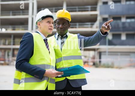 African-american and European men standing on construction site and talking. One of them holding folder with documentation, another one showing Stock Photo