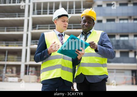 Engineer standing with builder supervising the construction process with house drawings on structure outdoors Stock Photo