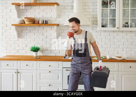 Man in overalls holding plunger like microphone singing while stands in modern kitchen with toolbox Stock Photo