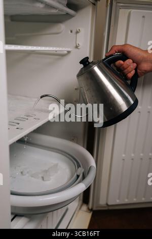 Housekeeper pouring hot water from kettle into refrigerator to melt ice and defrost, collecting melted ice in basin placed at bottom. Stock Photo
