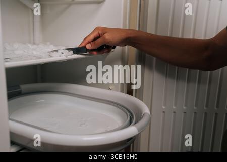 Person carefully defrosting their refrigerator, using knife to scrape off ice buildup and collecting ice chips in basin placed inside appliance for Stock Photo