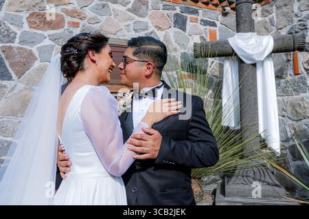 Bride in white dress caresses the cheek of groom in black tuxedo in front of a rustic stone wall and a large decorated cross. Stock Photo
