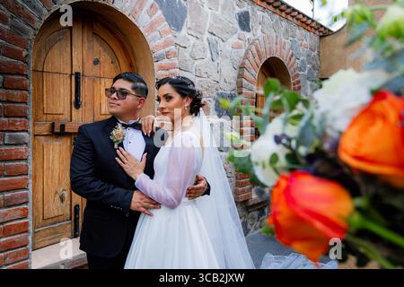 Bride and groom pose elegantly in front of stone wall and wooden gate, with vibrant bridal bouquet in the foreground. Stock Photo