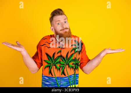 Young bearded man in tropical printed shirt gesturing casually against vibrant yellow background, expressing curiosity and humor Stock Photo