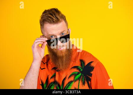 Casual red-bearded man in tropical print shirt adjusting sunglass on vibrant yellow background Stock Photo