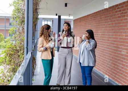 Diverse female coworkers holding white ceramic mugs and chatting on covered walkway outside office Stock Photo