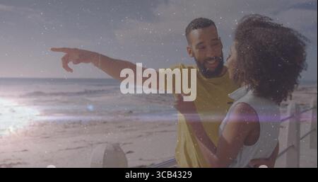 Smiling couple leaning on white railing pointing toward horizon and gazing on boardwalk, waves Stock Photo