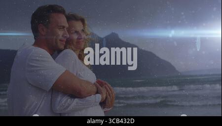Embracing middle-aged couple wearing white shirts watching dusk sky at beach with waves, copy space Stock Photo