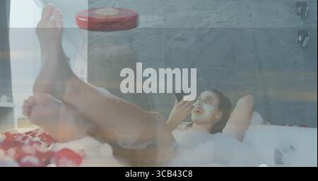 Holding smartphone woman relaxing in bubble bath at home, wearing white bikini top with rose petals Stock Photo