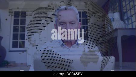 Sitting senior man wearing shirt using laptop on porch, with globe overlay, wicker chair and plant Stock Photo