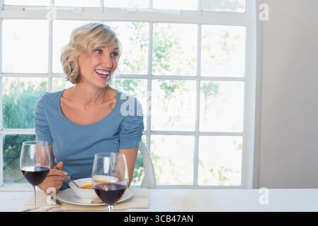 Middle-aged woman smiling while holding spoon over soup bowl with wine glasses at dining table Stock Photo