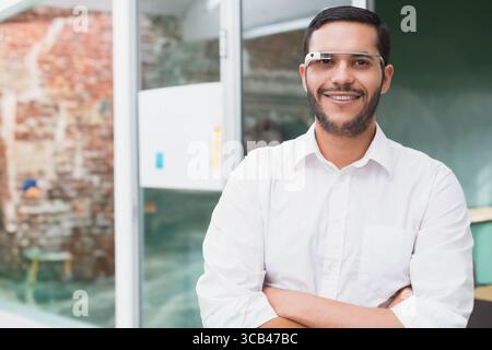 Man wearing white shirt and smart glasses with arms crossed at office showing green chalkboard Stock Photo