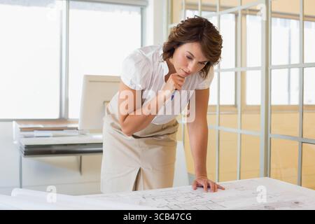 Woman wearing white blouse leaning over blueprints on table in office reviewing architectural plans Stock Photo