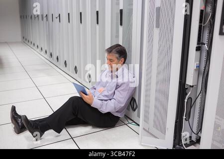 Middle-aged man IT technician sitting beside server rack in server room using tablet, copy space Stock Photo
