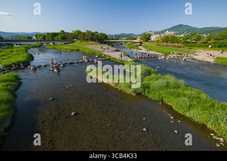 People enjoying a sunny day by a peaceful river in a green park at Kamogawa Delta. Families and friends gather on stepping stones and grassy areas, su Stock Photo