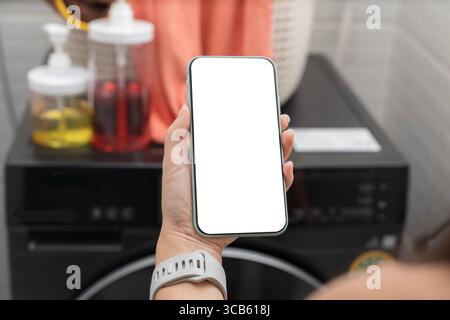 Hand holding smartphone with white screen for smart home or IoT app mockup. Background shows detergent bottles and washing machine. Concept of wireles Stock Photo